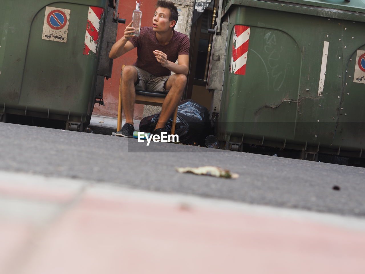 Surprised young man holding alcohol bottle amidst garbage cans