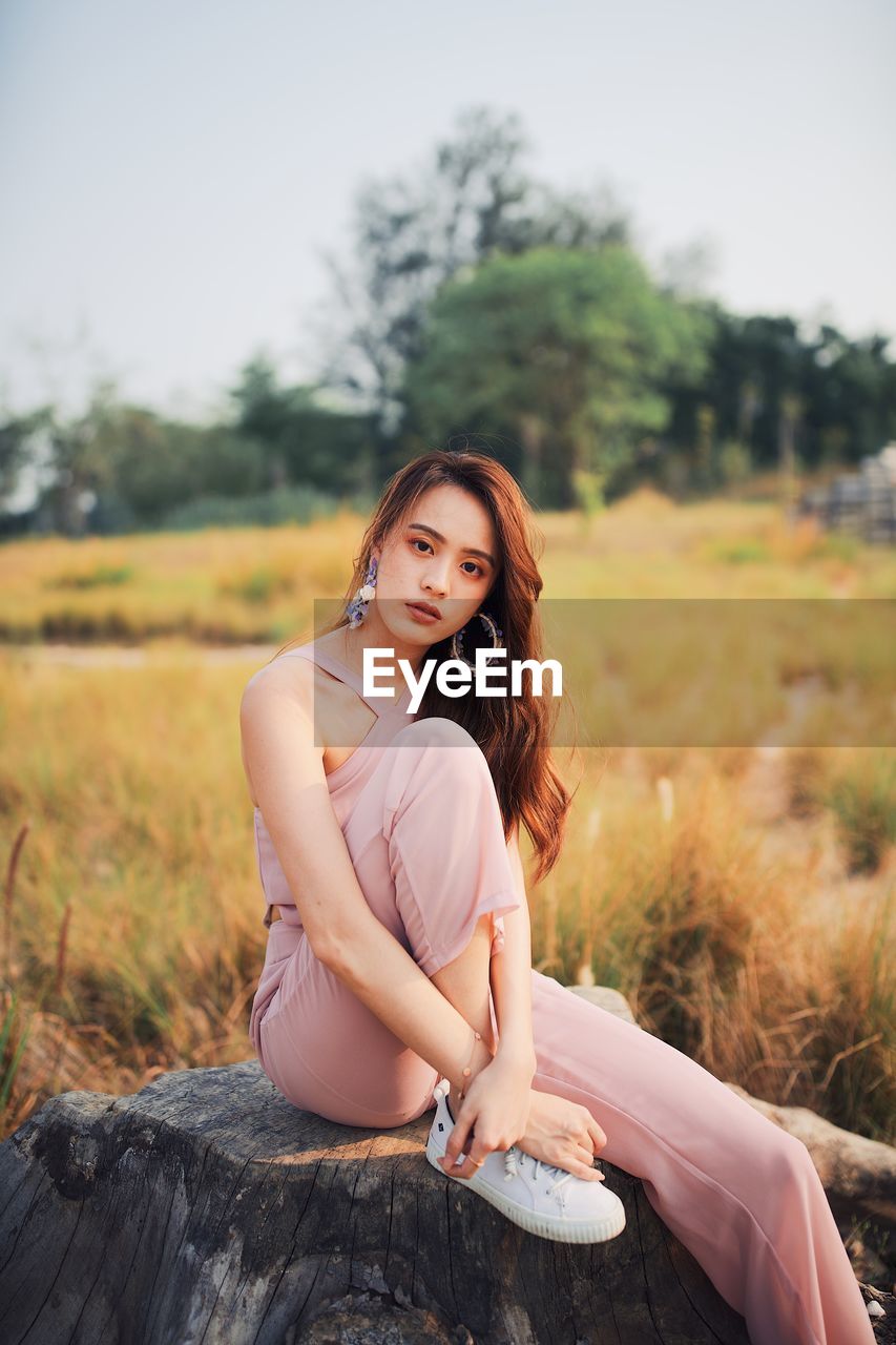 Portrait of young woman sitting on tree stump against clear sky