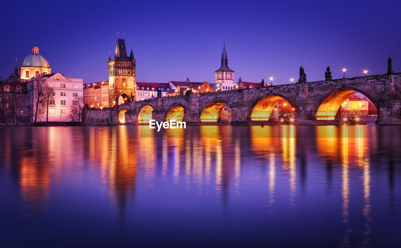 Charles bridge over river by buildings in illuminated city against clear sky