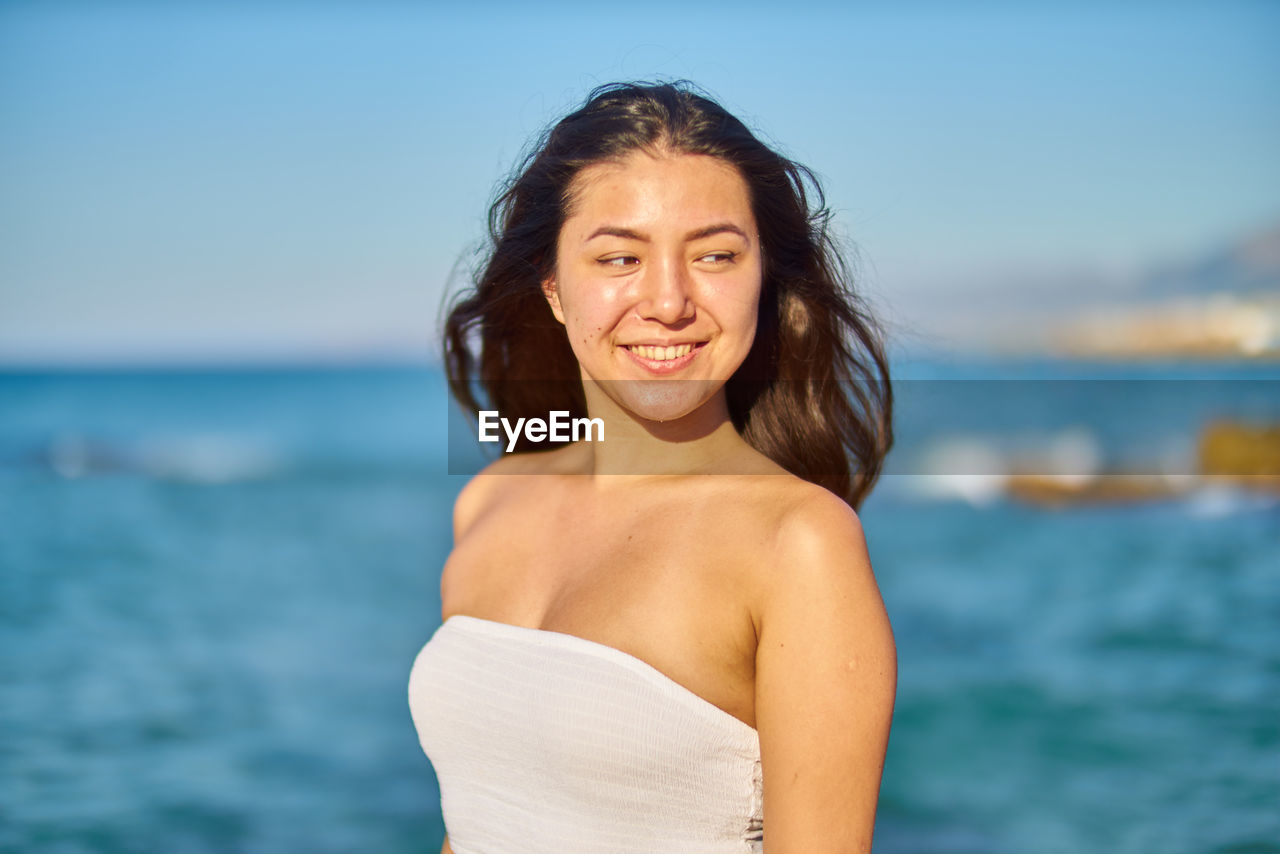 Portrait of smiling young woman standing at beach against sky