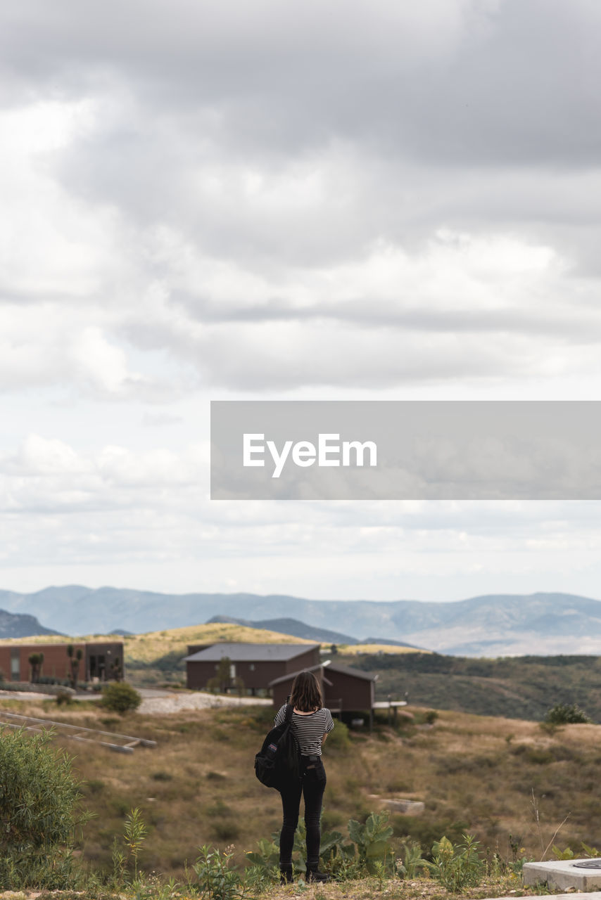 Rear view of woman standing on rock while looking at houses on mountain against sky