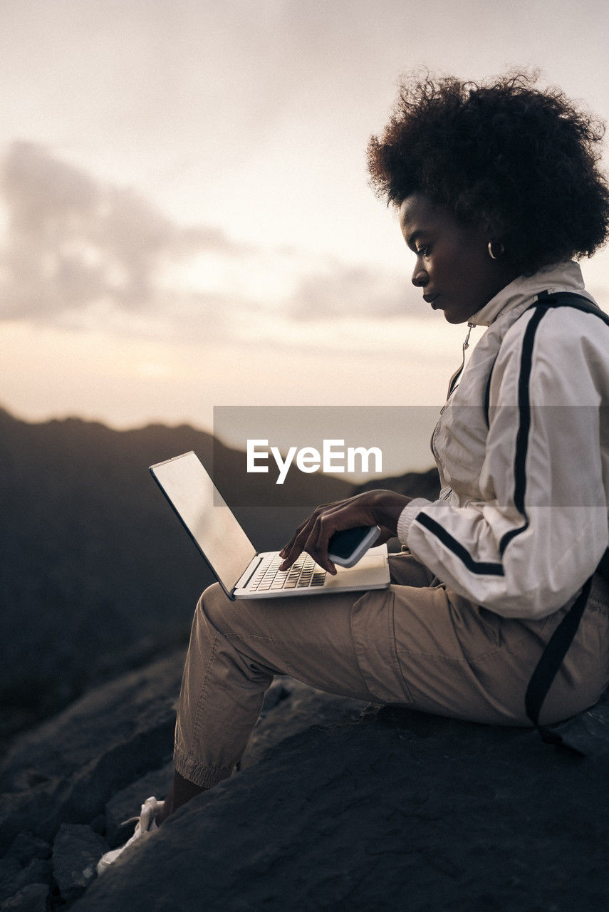 Woman with curly hair using laptop while sitting on rock at sunset