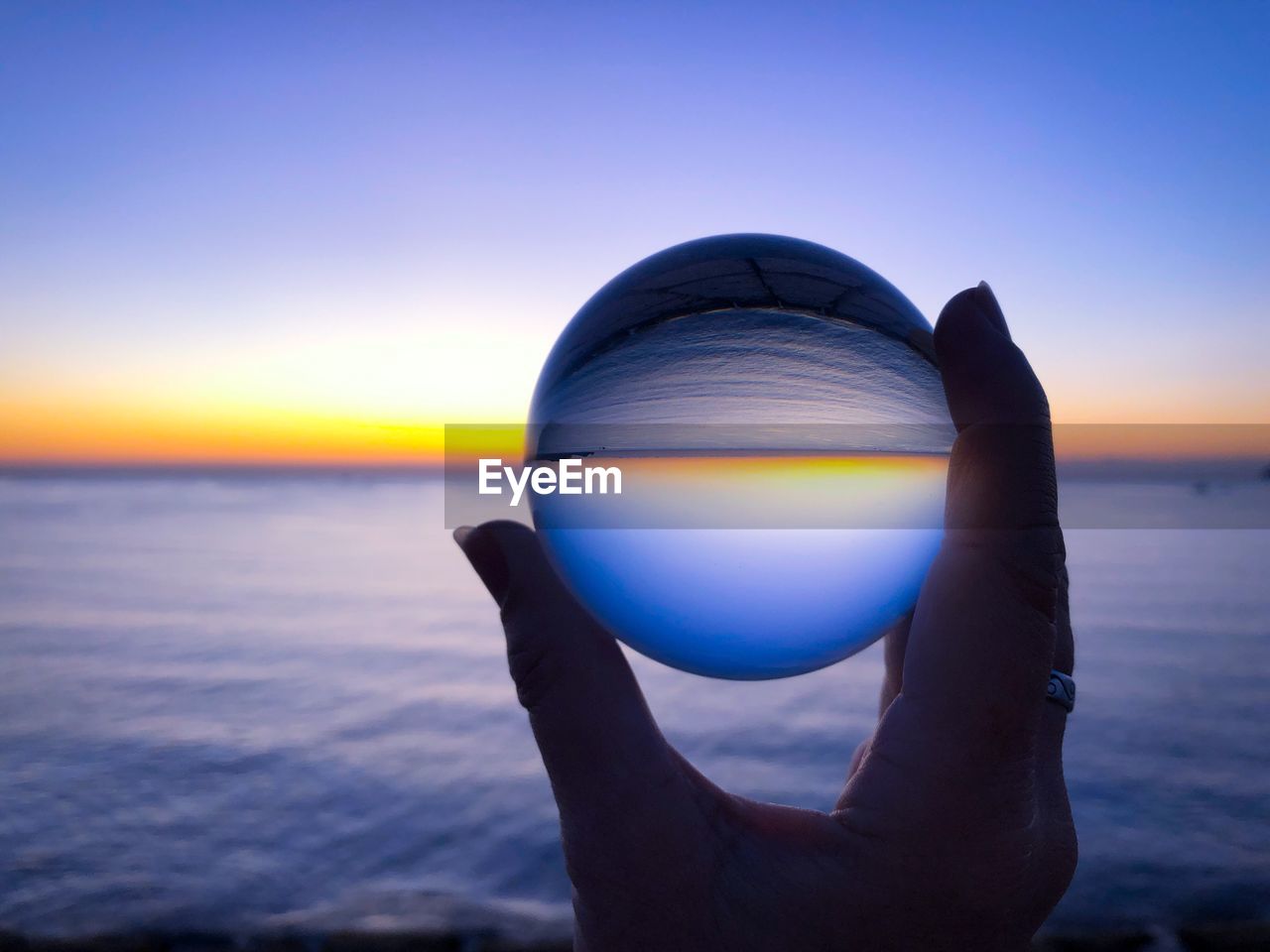 Cropped hand holding crystal ball against sky during sunset