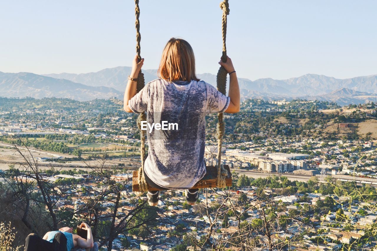 Rear view of woman sitting on swing against cityscape