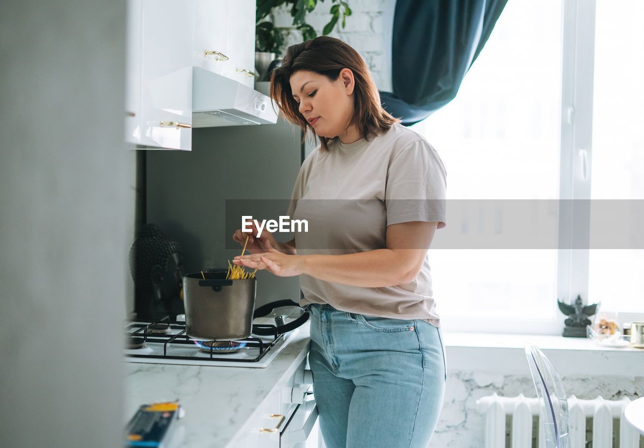 Beautiful brunette young woman plus size body positive cooking in kitchen at home