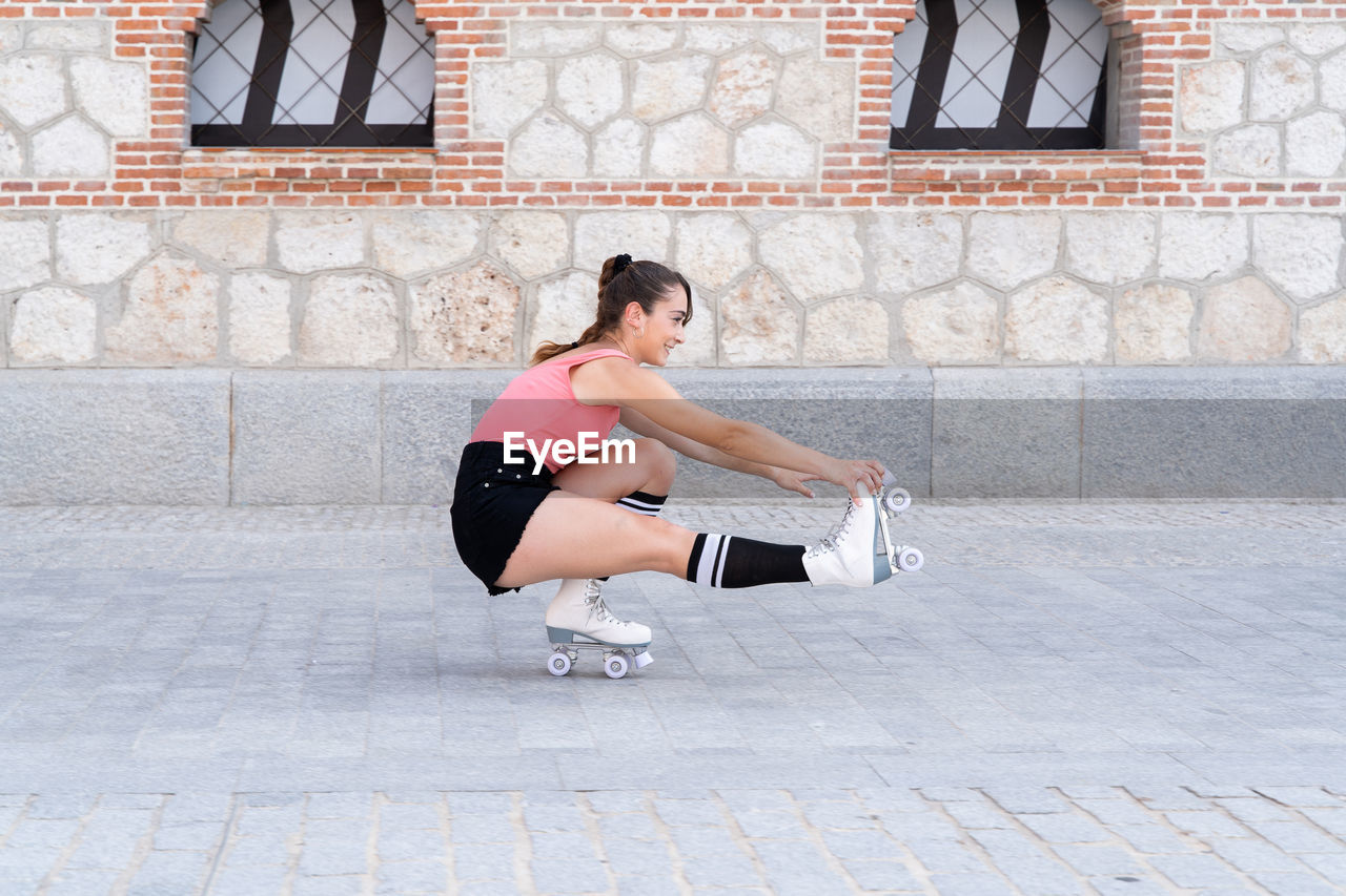 Full body side view of active hispanic female in roller skates doing stunt while riding on one leg on paved sidewalk near building