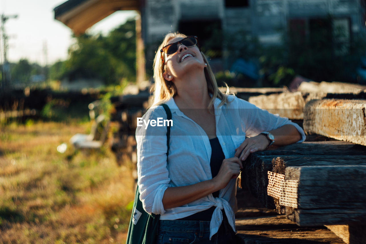 one person, adult, nature, women, focus on foreground, casual clothing, person, standing, sunlight, day, young adult, spring, three quarter length, lifestyles, tree, wood, clothing, architecture, hairstyle, outdoors, leisure activity, waist up, fashion, landscape, rural scene, emotion, glasses, sunglasses, female, long hair, built structure, plant, front view, blue, building exterior, looking, holding