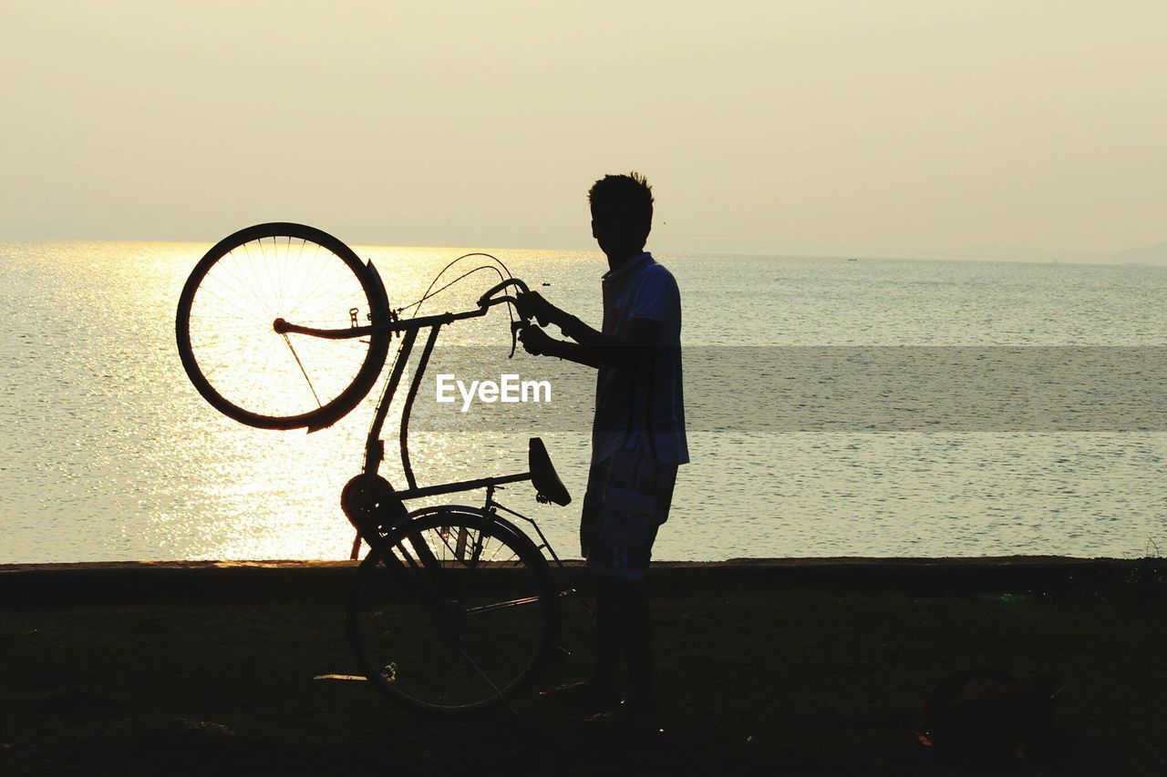 Silhouette man performing stunt with bicycle at beach against clear sky during sunset