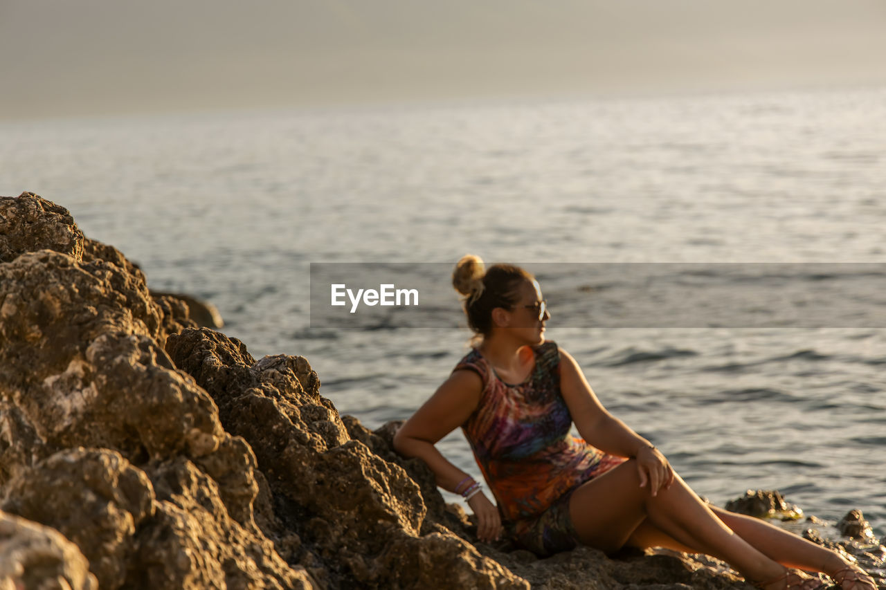 Side view of woman sitting at beach against sky during sunset