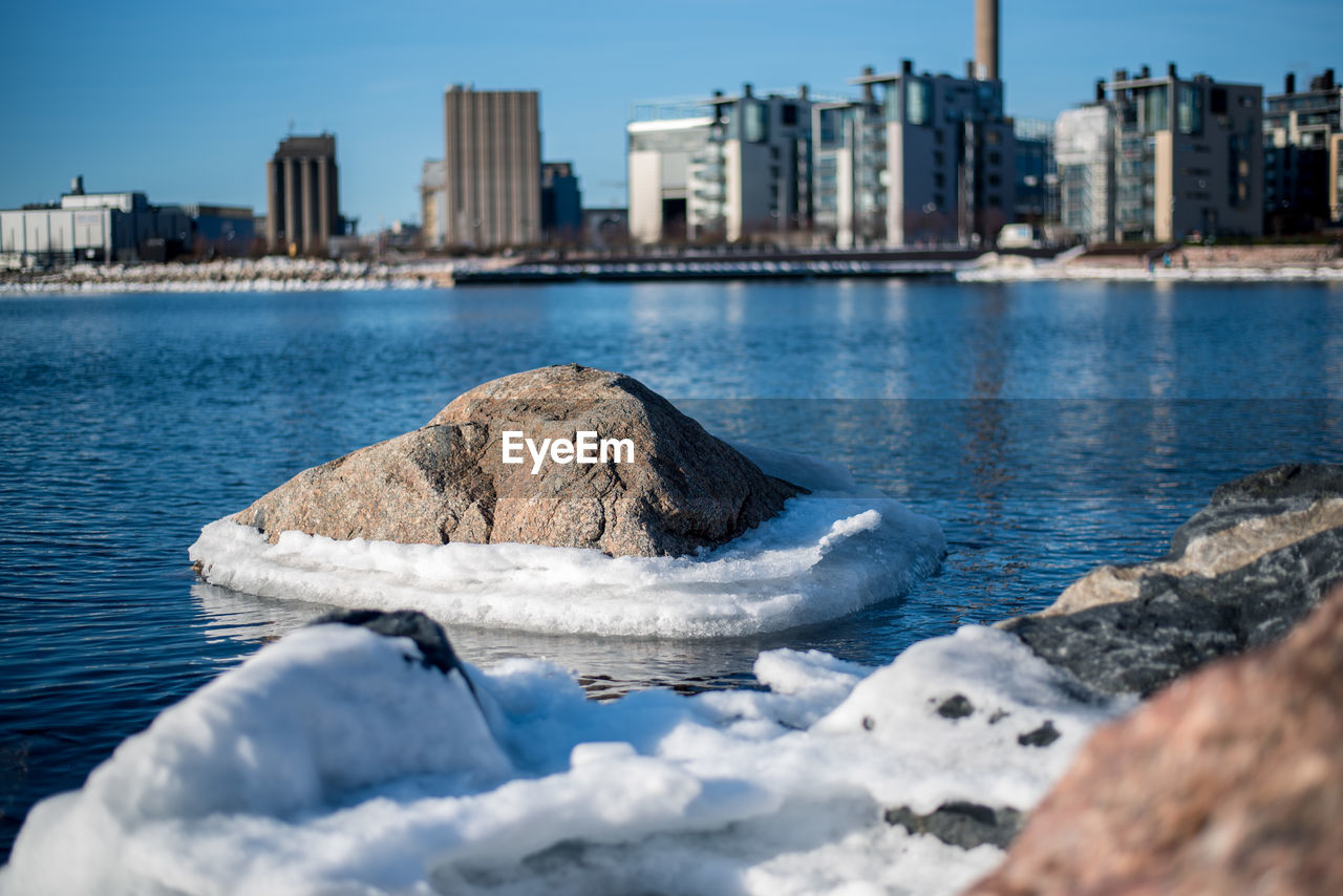 Rock by snow in river against skyscrapers