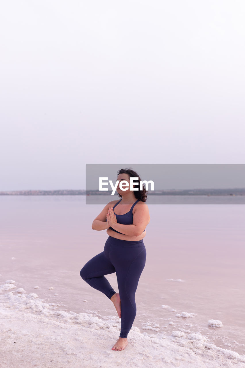 Serene curvy female in activewear standing in vrksasana on shore of pink lake and practicing yoga