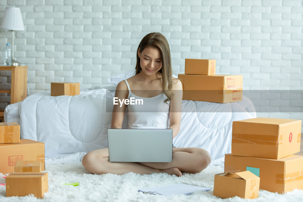 Full length of woman using laptop while sitting amidst cardboard boxes at home