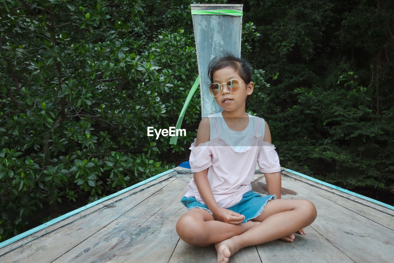 Full length of girl wearing sunglasses sitting on boat deck against tree