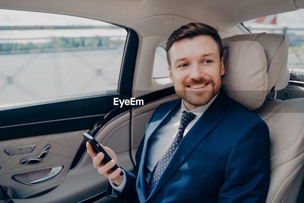 Smiling businessman holding mobile phone sitting at car