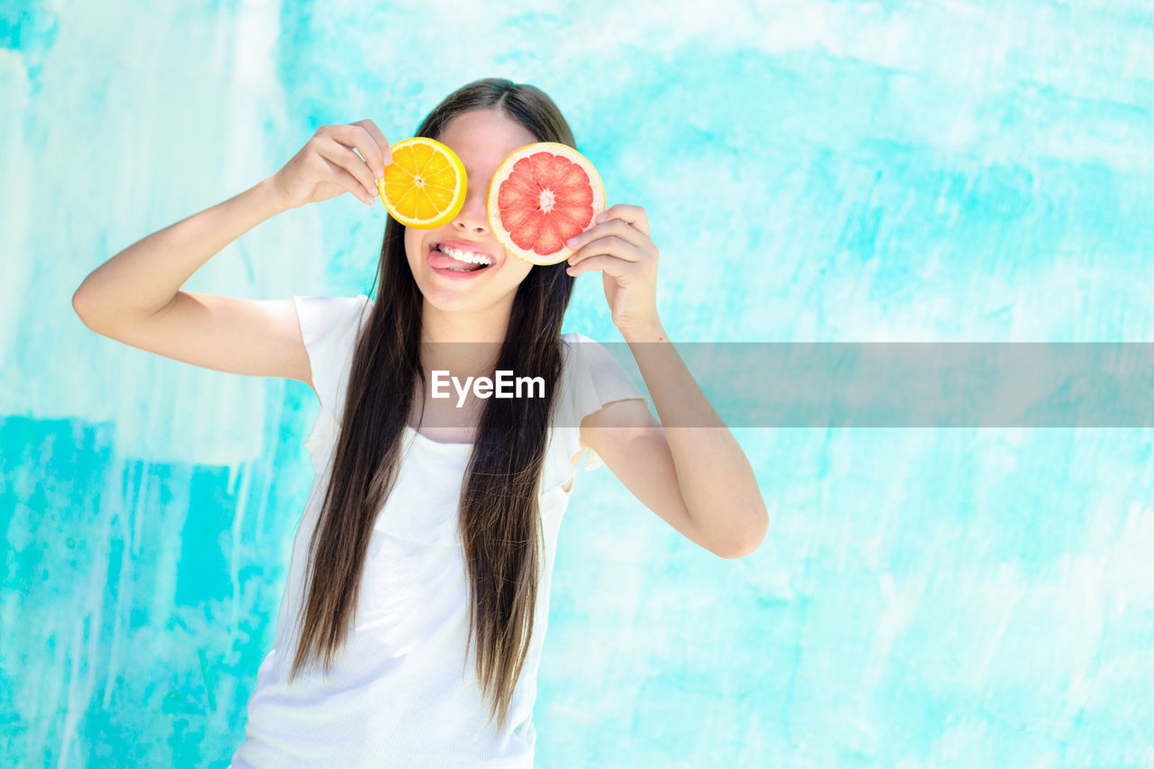 Happy teenage girl holding citrus fruit slices in front of eyes while standing by turquoise wall