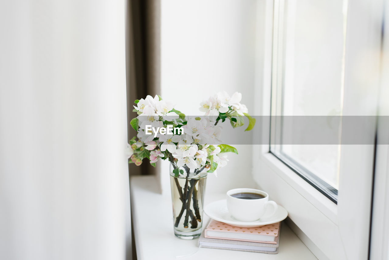 White cup of coffee on a white plate, a stack of books and a vase of flowers on the windowsill 