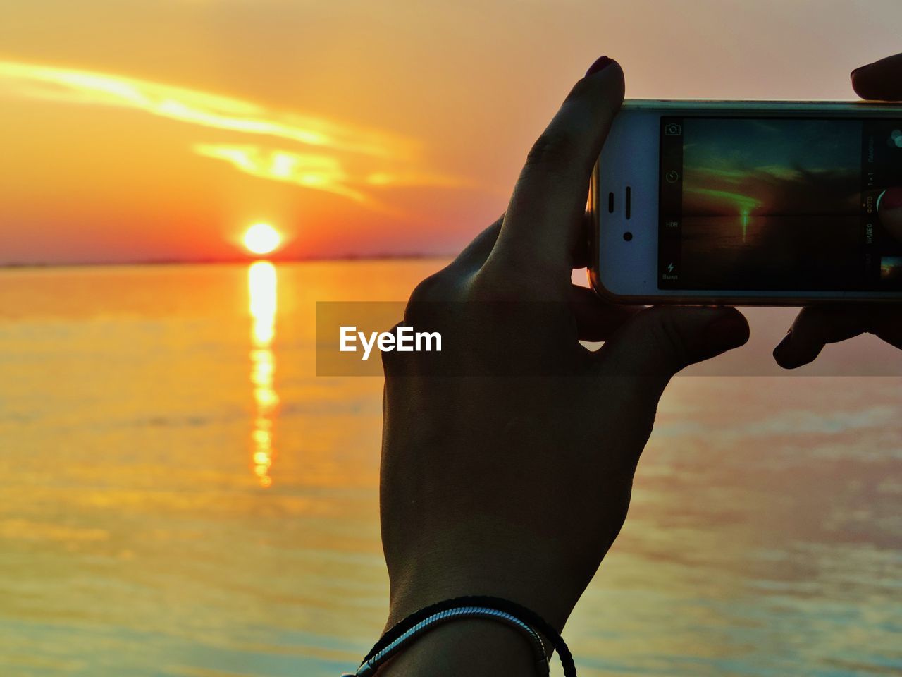 HUMAN HAND HOLDING CAMERA AGAINST SEA DURING SUNSET