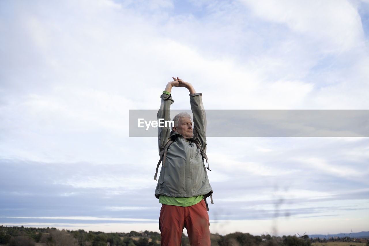 Senior male hiker stretching arms while standing against cloudy sky at countryside