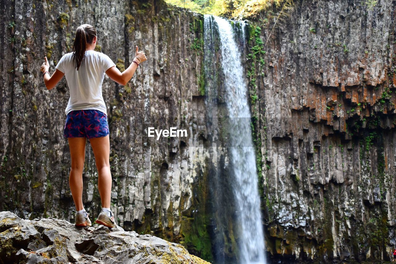 Rear view of woman gesturing thumbs up on rock near waterfall