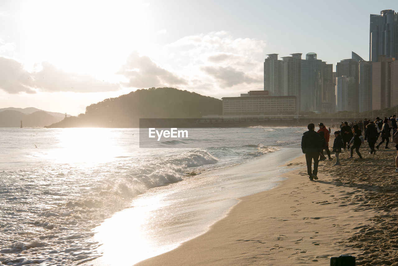 PEOPLE ON BEACH BY SEA AGAINST SKY DURING SUNSET