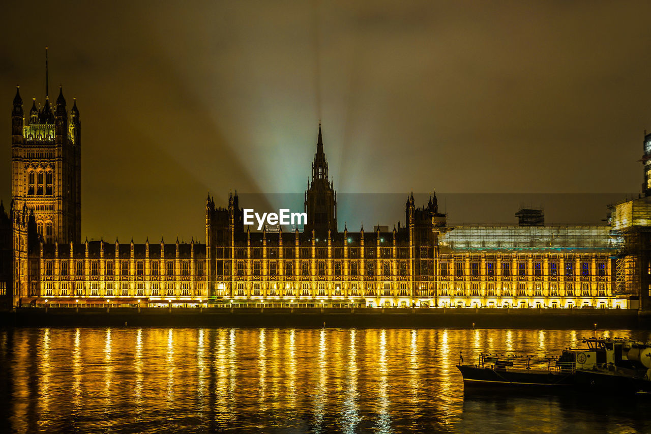 illuminated buildings by river against sky at night