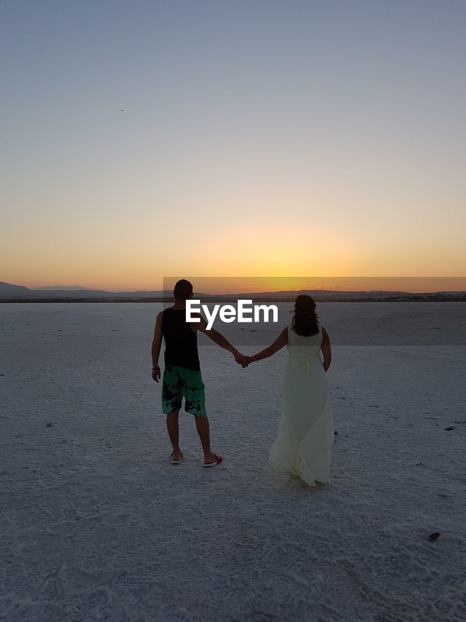 Rear view of couple holding hands walking at beach against sky during sunset