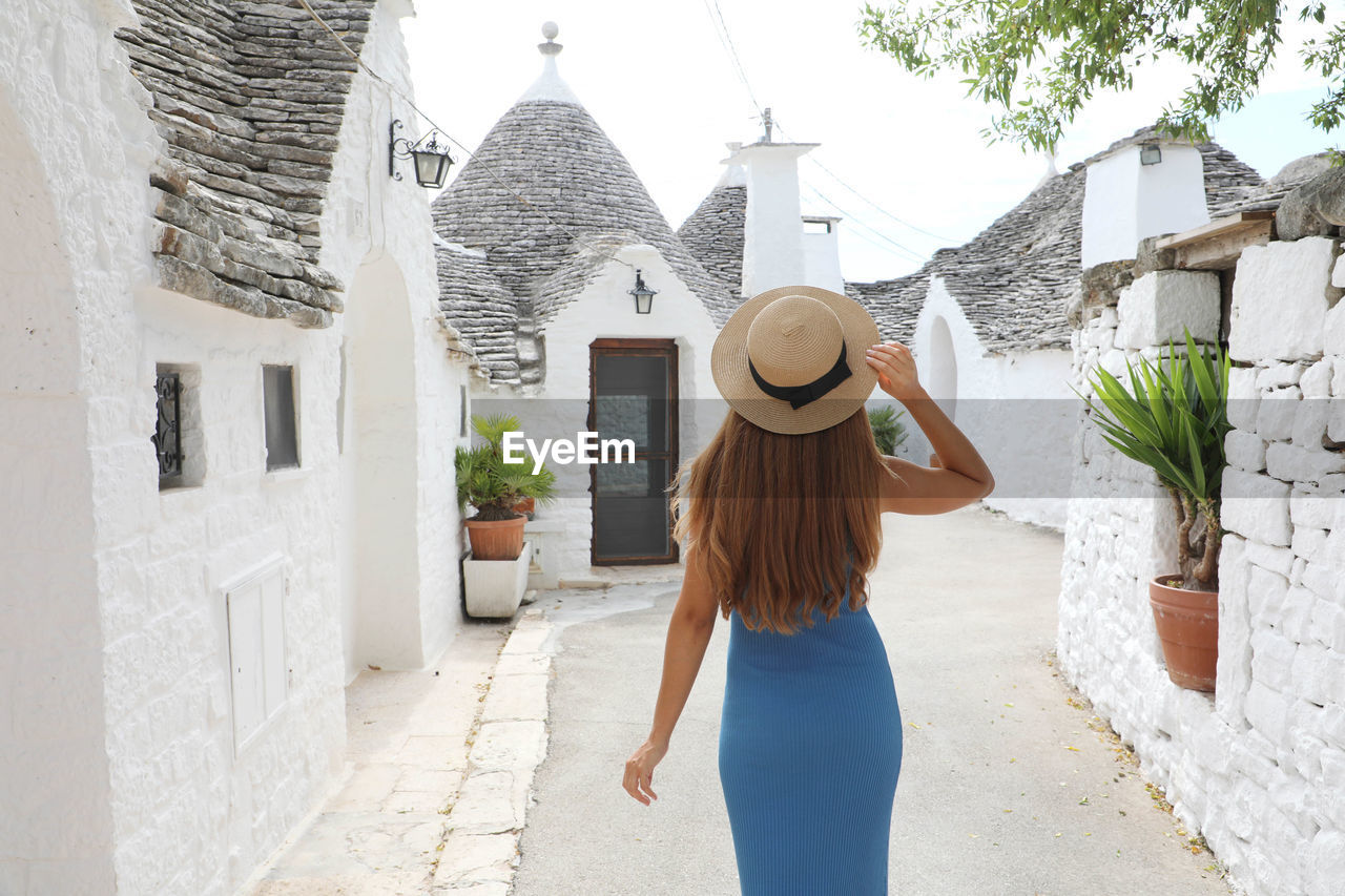 Curious young woman with blue dress and hat walking in street of alberobello, italy. 