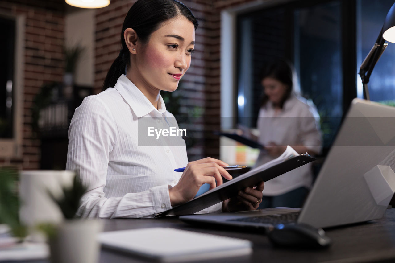 Young businesswoman holding file folder looking at laptop screen in office