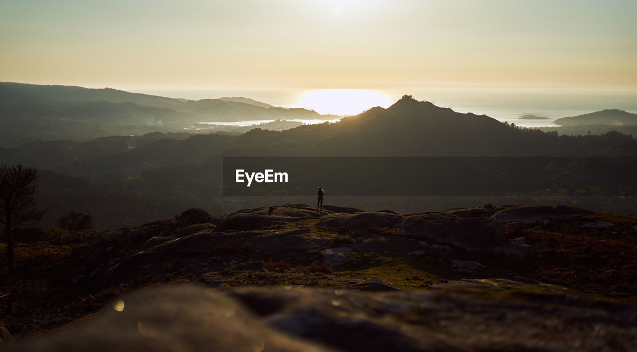 Distant view of woman standing against mountain and sky during sunset