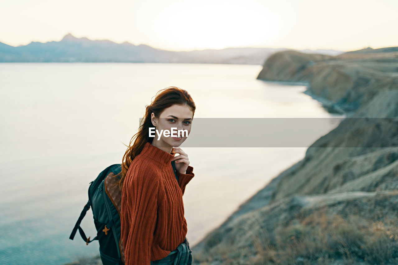 PORTRAIT OF SMILING YOUNG WOMAN STANDING BY SEA