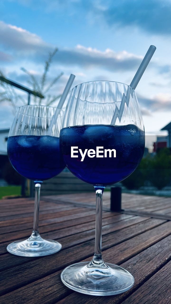 blue, food and drink, glass, refreshment, drink, alcohol, cloud, sky, wine glass, drinking glass, stemware, household equipment, wood, nature, no people, wine, cocktail, food, table, freshness, focus on foreground, outdoors, reflection, water, day, alcoholic beverage, tableware