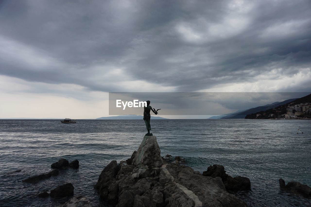 Man standing on rock at beach against cloudy sky