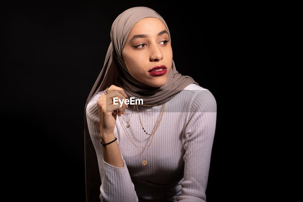 Modish confident muslim female in hijab leaning on chair and looking away in dark studio