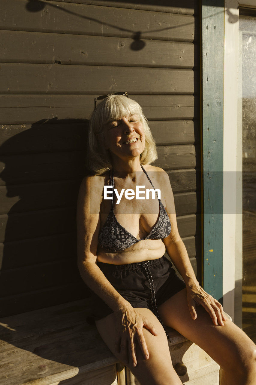 Smiling elderly woman sunbathing with eyes closed while sitting on houseboat