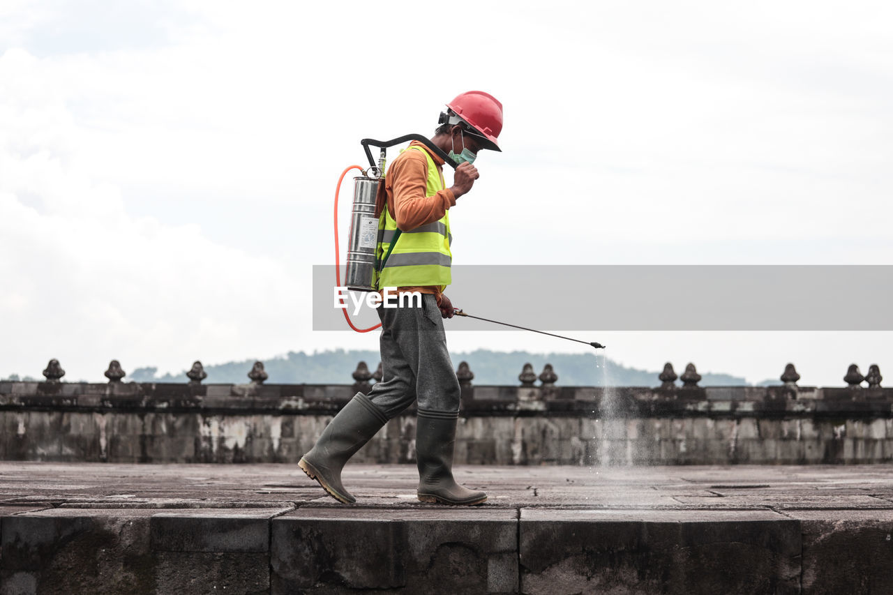 Side view of worker spraying chemical on footpath against sky