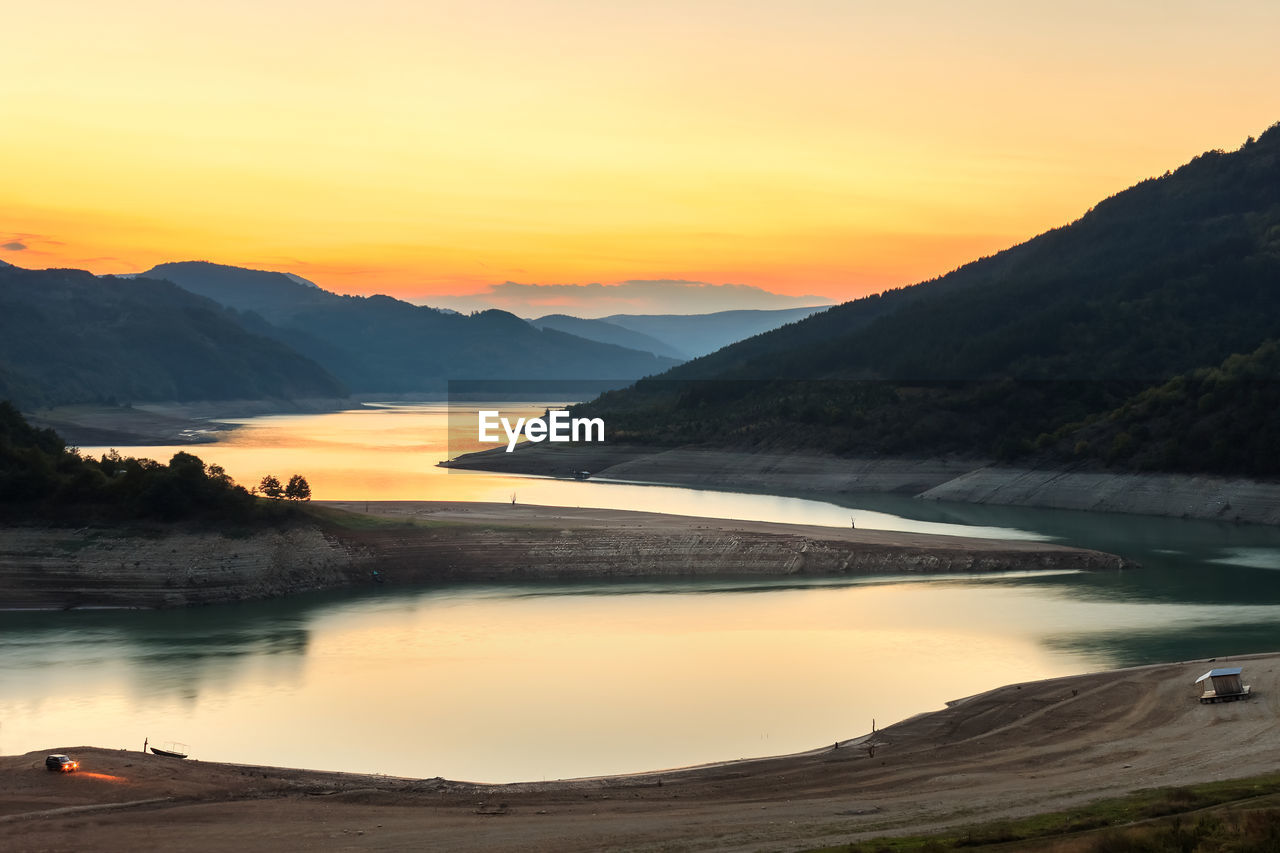 SCENIC VIEW OF LAKE BY MOUNTAINS AGAINST ORANGE SKY