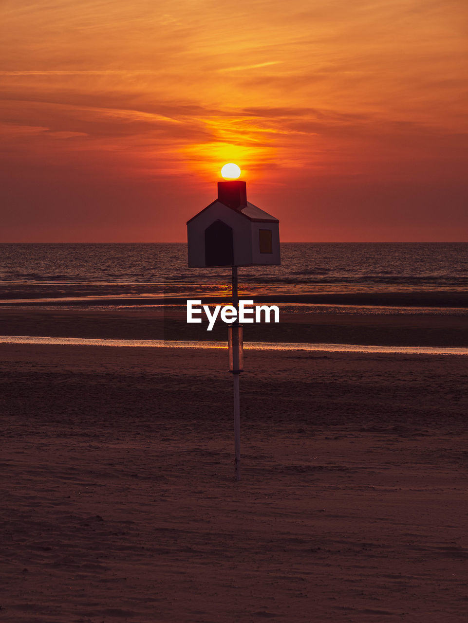 Silhouette of a wooden house on the beach with the sun sitting on the chimney during sunset