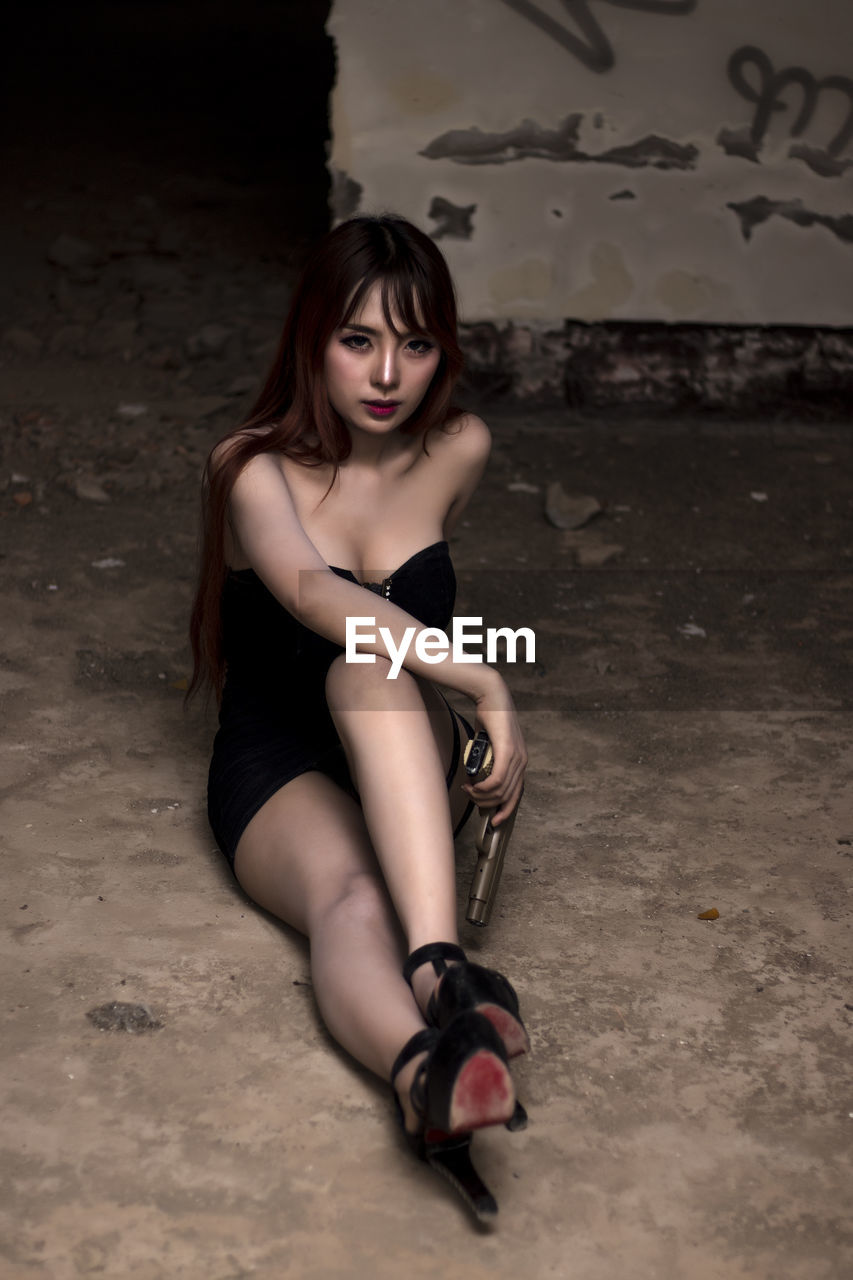 Portrait of beautiful woman holding handgun while sitting on floor in abandoned room