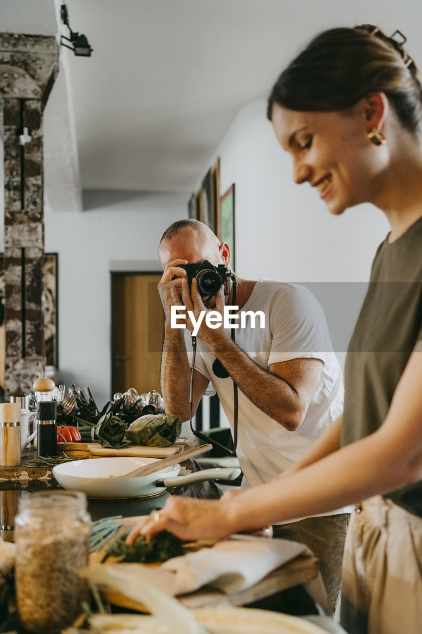 Colleague photographing female chef through camera while preparing food in studio