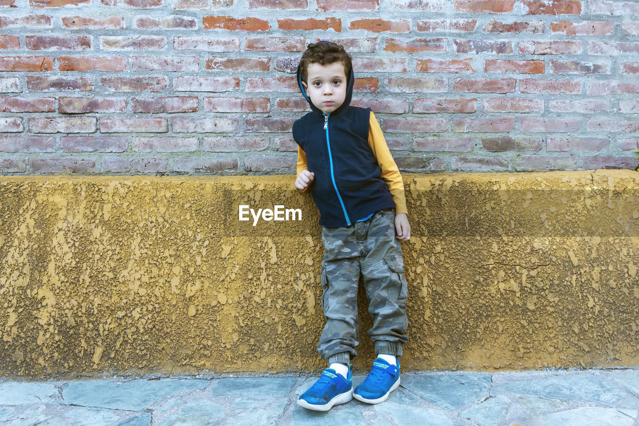 Boy in hooded sweatshirt and camouflage pants leaning against brick wall