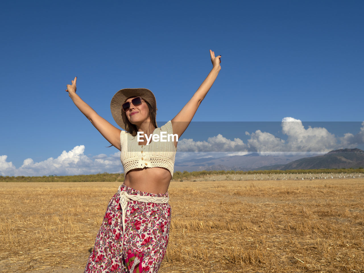 sky, one person, adult, women, arm, landscape, fashion, nature, happiness, land, limb, arms raised, smiling, emotion, human limb, environment, blue, clothing, cheerful, summer, rural scene, young adult, sunlight, positive emotion, glasses, standing, field, portrait, beauty in nature, sunglasses, day, joy, sunny, enjoyment, cloud, travel, copy space, female, fun, outdoors, arms outstretched, vacation, scenics - nature, trip, leisure activity, carefree, front view, holiday, person, excitement, looking at camera, vitality, lifestyles, hand, hand raised, agriculture, photo shoot, travel destinations, plant, success, horizon, dress