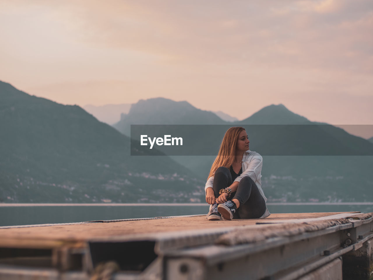 WOMAN SITTING ON MOUNTAINS AGAINST SKY DURING SUNSET