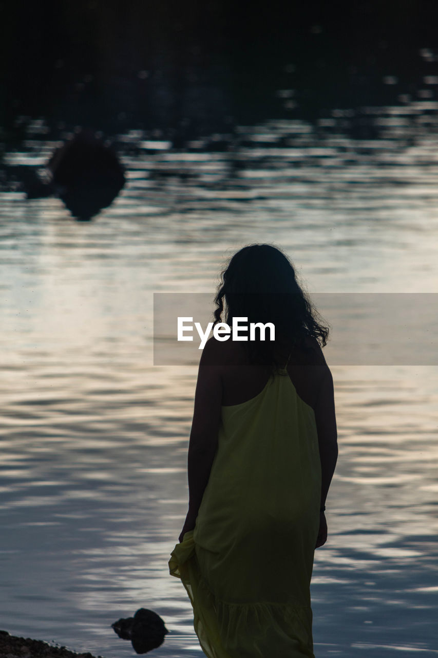 REAR VIEW OF WOMAN STANDING IN LAKE AT SUNSET