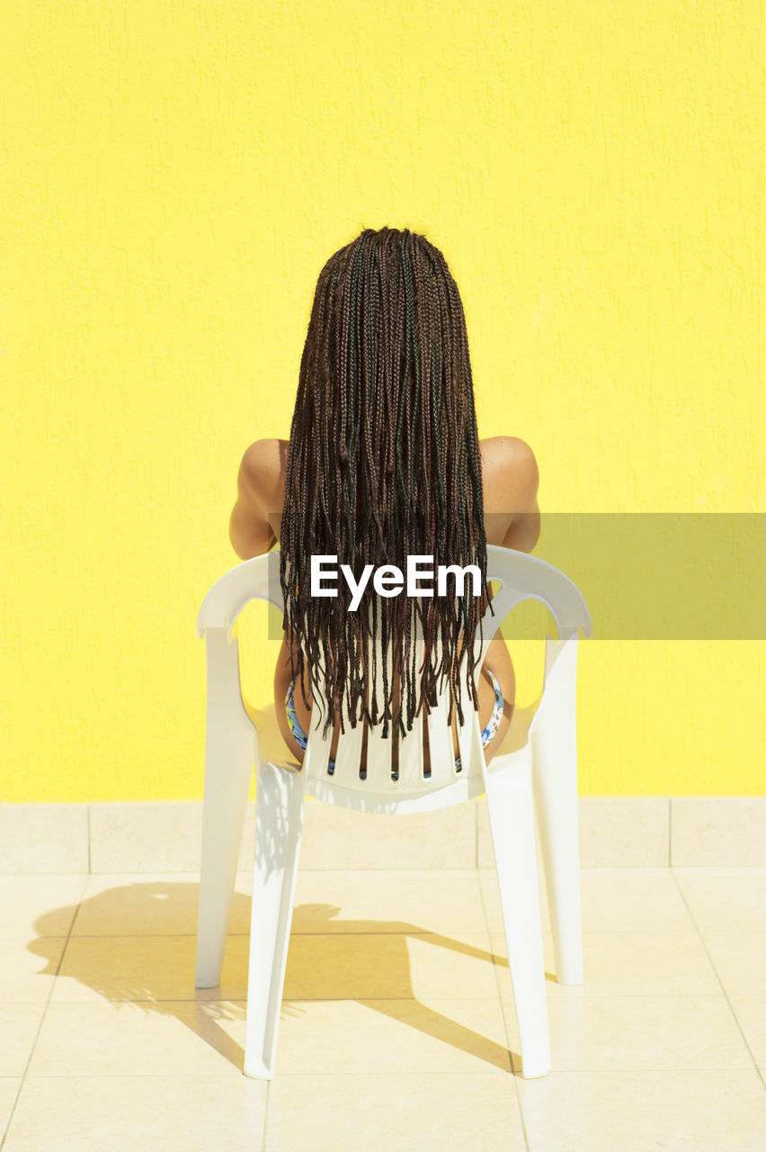 Rear view of woman sitting on chair against yellow wall