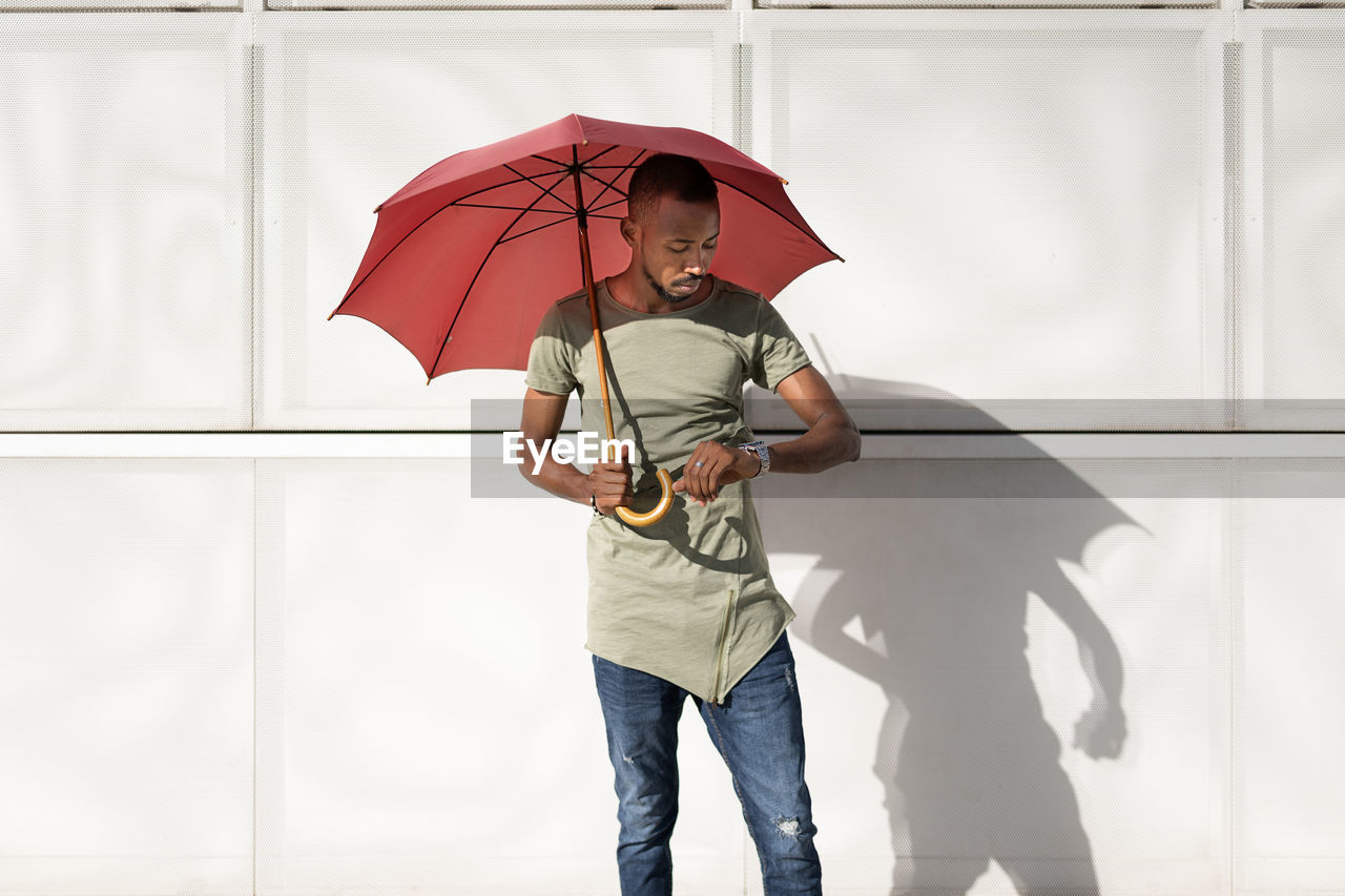 Serious african american male standing under umbrella on street on sunny day and checking time on wristwatch