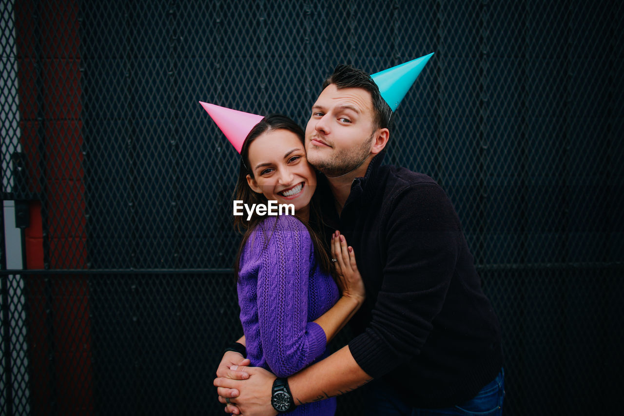 Portrait of man embracing woman while wearing party hats against metal grate