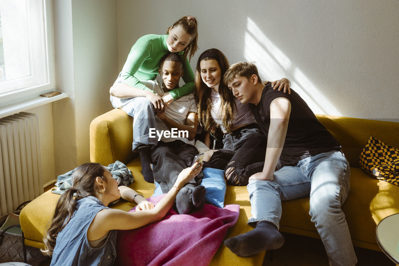 Young woman sharing smart phone with multiracial friends sitting together on sofa at home