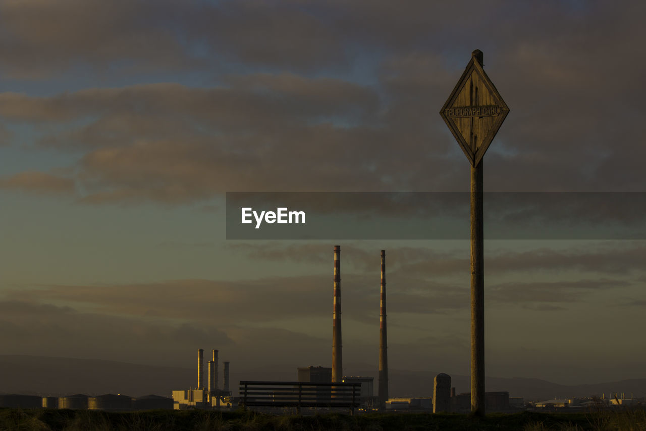 Pole and smoke stacks against cloudy sky at sunset