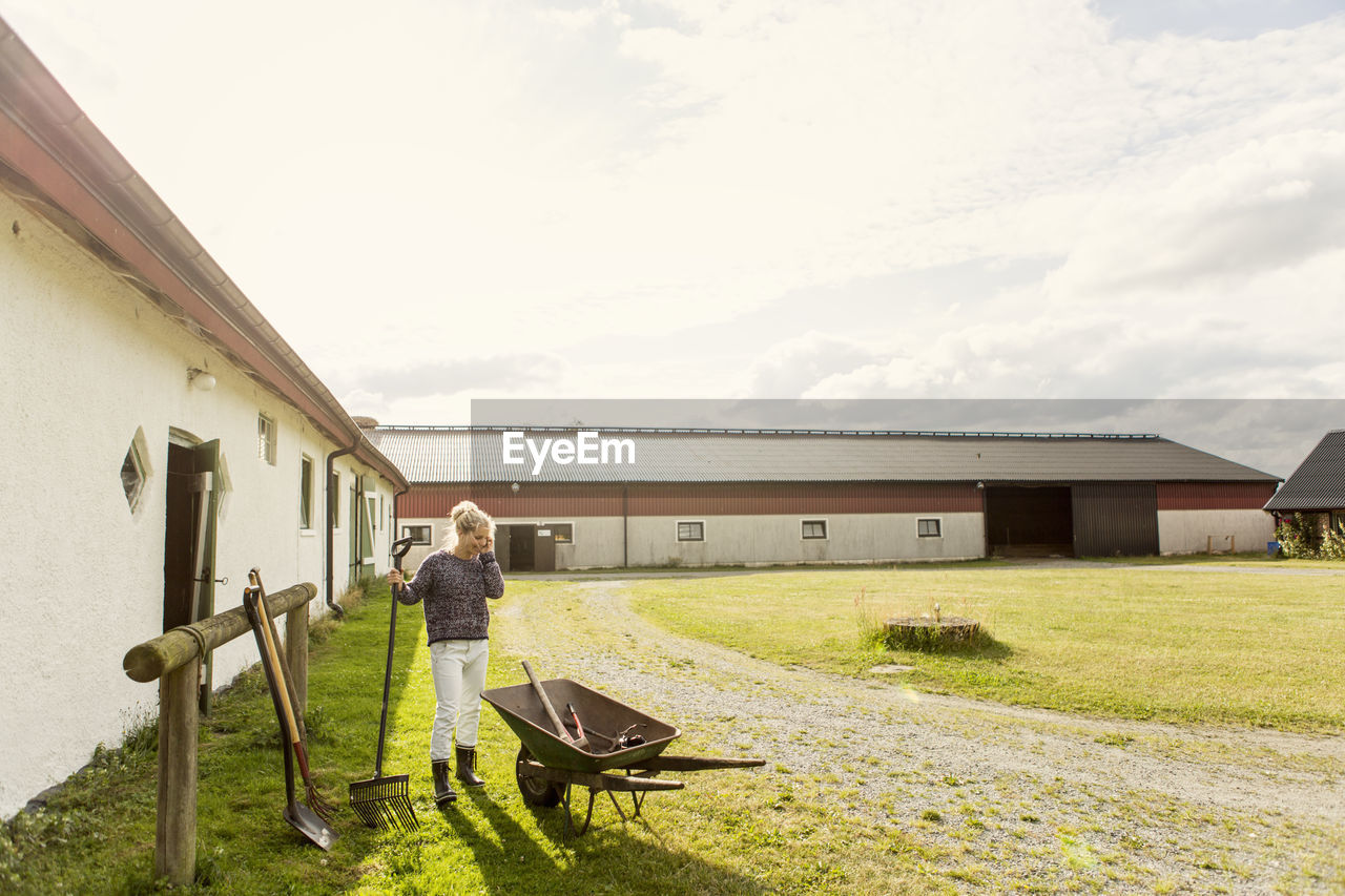 Woman using phone while holding rake against barn at farm