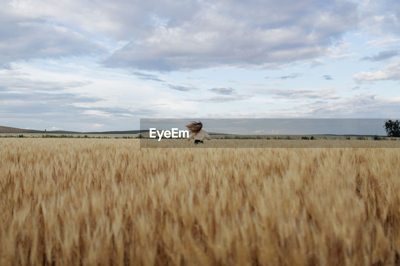 Back view of anonymous female with flying hair running on meadow with wheat spikes under cloudy sky